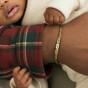 May include: A gold-colored chain bracelet with three rectangular charms, each engraved with a name: Chloe, Anabella, and Scarlett. The bracelet is worn on a wrist, with a baby's hand and face visible in the background. The bracelet is a personalized jewelry item.