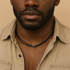 Può includere: Una collana nera con una piccola perla cilindrica color argento. La collana è composta da due fili di cordoncino nero. La persona nell'immagine indossa una camicia color beige.