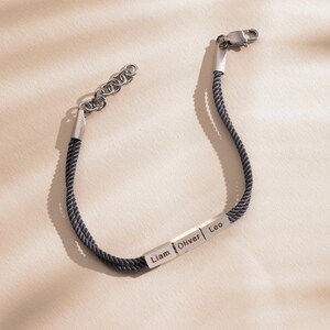 May include: A black braided cord bracelet with three silver rectangular charms engraved with the names Liam, Oliver, and Leo.