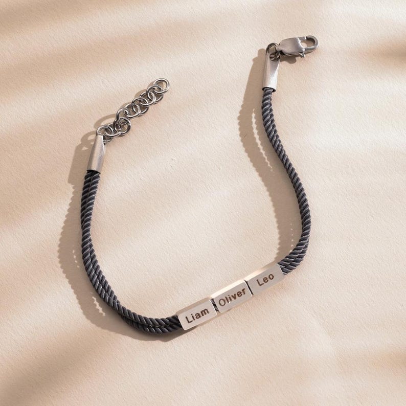 May include: A black braided cord bracelet with three silver rectangular charms engraved with the names Liam, Oliver, and Leo.