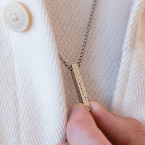 Puede incluir: Un collar de cadena de plata con un colgante de barra de plata grabado con puntos Braille.