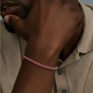 May include: A pink and white braided bracelet is shown on a wrist. The bracelet is made of a woven material. The person is wearing a beige collared shirt.