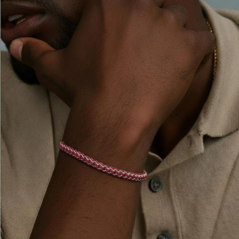 May include: A pink and white braided bracelet is shown on a wrist. The bracelet is made of a woven material. The person is wearing a beige collared shirt.
