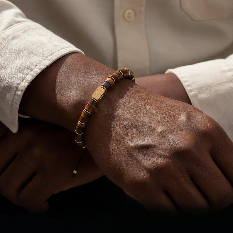 May include: A beaded bracelet featuring tiger's eye stones and gold-colored accents. The bracelet has a rectangular gold-colored center piece. The bracelet is on a black cord with an adjustable knot.