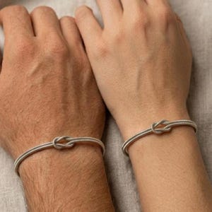 May include: Two silver-toned bracelets, each featuring a knot design at the center. The bracelets are worn on wrists, with the knot detail prominently displayed. The bands have a textured appearance, and the overall aesthetic is minimalist and elegant.