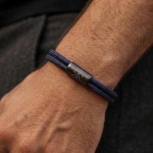 Könnte beinhalten: Ein dunkelblaues Armband aus zwei Kordelsträngen mit einer zylindrischen, mattschwarzen Metallperle. Die Perle ist mit einem kleinen Herzsymbol über einem X-förmigen Design graviert, wobei die Buchstaben L und P links und T rechts stehen.