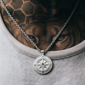 May include: Silver-toned compass pendant necklace. The round pendant features a detailed compass rose design with directional markers. The necklace is worn on a person wearing a gray ribbed tank top. The chain is a box chain style.