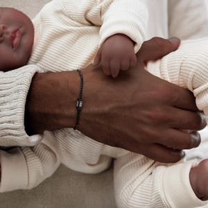 Puede incluir: Una pulsera trenzada negra con un dije plateado que dice "Liam". La pulsera se lleva en la muñeca de una persona que sostiene un bebé.