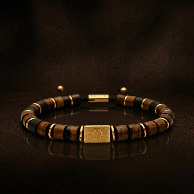 May include: A beaded bracelet featuring alternating brown and black wooden beads with gold-colored accents. The bracelet includes a gold-colored rectangular centerpiece with a horseshoe symbol. The bracelet is on a dark brown surface.