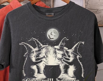May include: Dark gray t-shirt featuring a whimsical illustration of three cats in witch hats gathered around a cauldron under a full moon. The text "Cats will be cats" and "Est. 9 lives ago" are printed below the image. Other clothing hangs in the background.