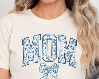 May include: A white t-shirt with a blue floral print of the word "MOM" in a collegiate font with a blue bow underneath.