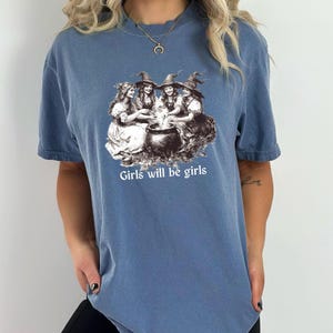以下が含まれることがあります： 3人の魔女が釜の周りにいるグラフィックがプリントされた青いTシャツ。グラフィックの下に「Girls will be girls」というテキストがプリントされています。