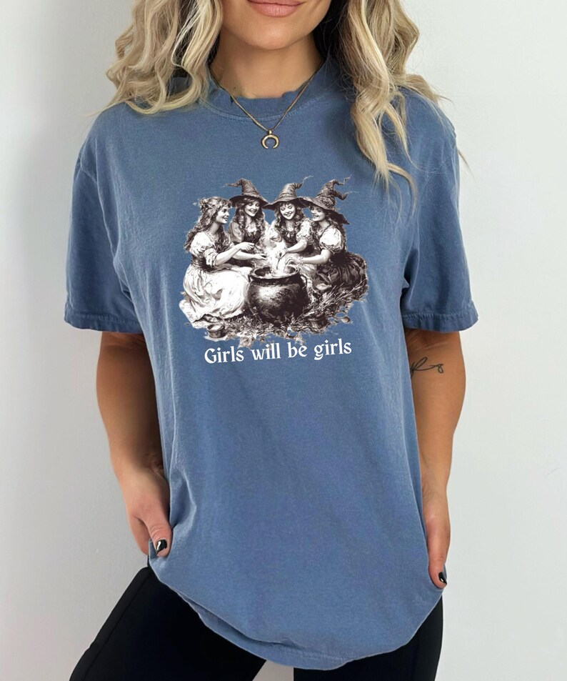 以下が含まれることがあります： 3人の魔女が釜の周りにいるグラフィックがプリントされた青いTシャツ。グラフィックの下に「Girls will be girls」というテキストがプリントされています。