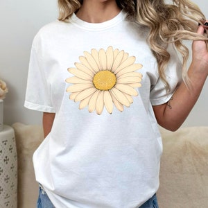 Puede incluir: Camiseta blanca con un gran gr&aacute;fico de margarita de color crema. La margarita tiene un centro amarillo y est&aacute; centrada en la camiseta.