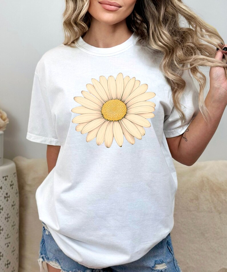 Puede incluir: Camiseta blanca con un gran gr&aacute;fico de margarita de color crema. La margarita tiene un centro amarillo y est&aacute; centrada en la camiseta.
