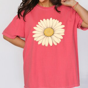 Puede incluir: Una camiseta de gran tama&ntilde;o de color rosa coral con un gr&aacute;fico de margarita blanca.