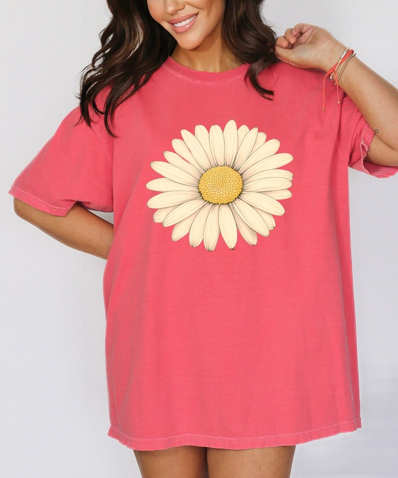 Puede incluir: Una camiseta de gran tama&ntilde;o de color rosa coral con un gr&aacute;fico de margarita blanca.