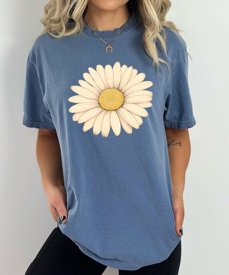 Puede incluir: Una camiseta azul con un gr&aacute;fico de margarita blanca. La margarita tiene un centro amarillo.
