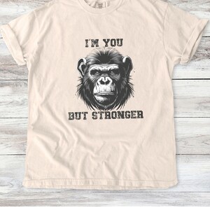 Peut inclure: Un t-shirt beige avec une illustration en noir et blanc d'un visage de singe et le texte "I'm you but stronger".