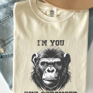 Peut inclure: Un t-shirt beige avec une illustration en noir et blanc d'un visage de singe. Le texte "I'm you but stronger" est imprim&eacute; sous l'image.