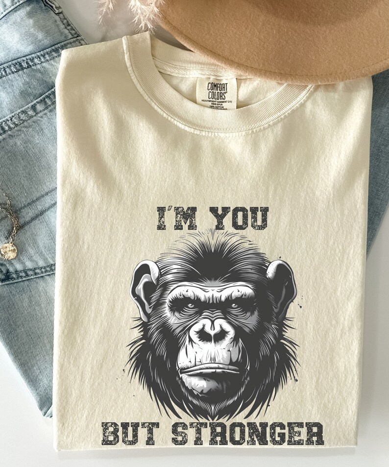 Peut inclure: Un t-shirt beige avec une illustration en noir et blanc d'un visage de singe. Le texte "I'm you but stronger" est imprim&eacute; sous l'image.