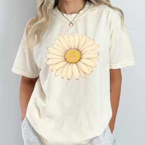 Puede incluir: Una camiseta de color crema con un gran gr&aacute;fico de margarita blanca. La margarita tiene un centro amarillo.