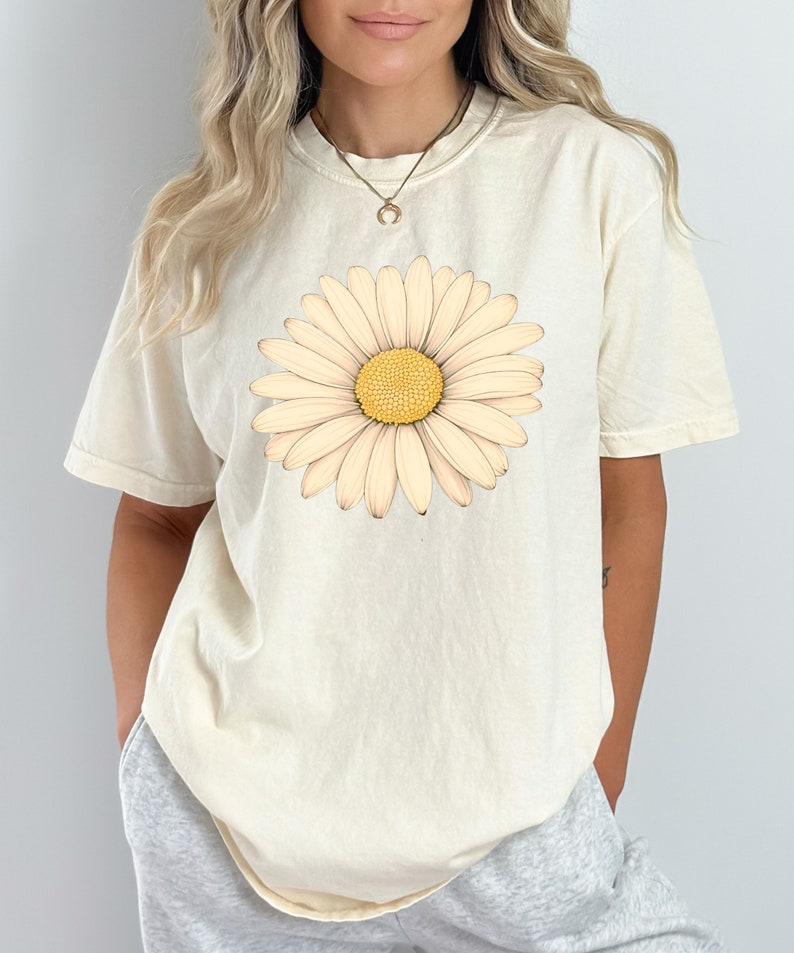 Puede incluir: Una camiseta de color crema con un gran gr&aacute;fico de margarita blanca. La margarita tiene un centro amarillo.