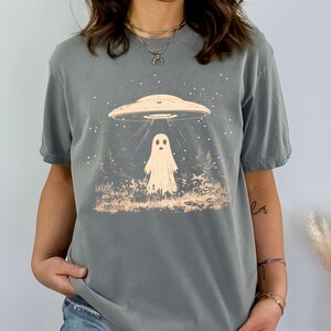 Puede incluir: Una camiseta gris con un gr&aacute;fico de un fantasma siendo abducido por un OVNI. El gr&aacute;fico es de estilo vintage con un fondo de noche estrellada.