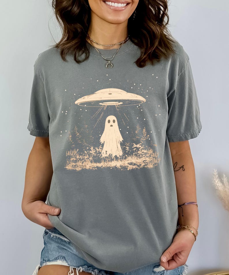 Puede incluir: Una camiseta gris con un gr&aacute;fico de un fantasma siendo abducido por un OVNI. El gr&aacute;fico es de estilo vintage con un fondo de noche estrellada.
