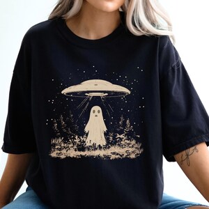 Puede incluir: Una camiseta negra con un estampado gr&aacute;fico de un fantasma de pie frente a un OVNI. El gr&aacute;fico est&aacute; en un estilo vintage con un color marr&oacute;n claro.