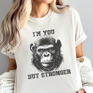 Peut inclure: Un t-shirt blanc avec une illustration en noir et blanc d'un visage de singe. Le texte "I'M YOU BUT STRONGER" est imprim&eacute; sous l'image.