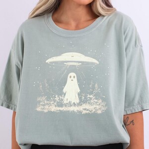 Puede incluir: Una camiseta verde claro con un gr&aacute;fico de un fantasma de pie en un bosque con un OVNI flotando arriba. El gr&aacute;fico es blanco.