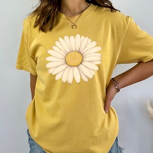 Puede incluir: Una camiseta amarilla de manga corta con un estampado gr&aacute;fico de margarita blanca.