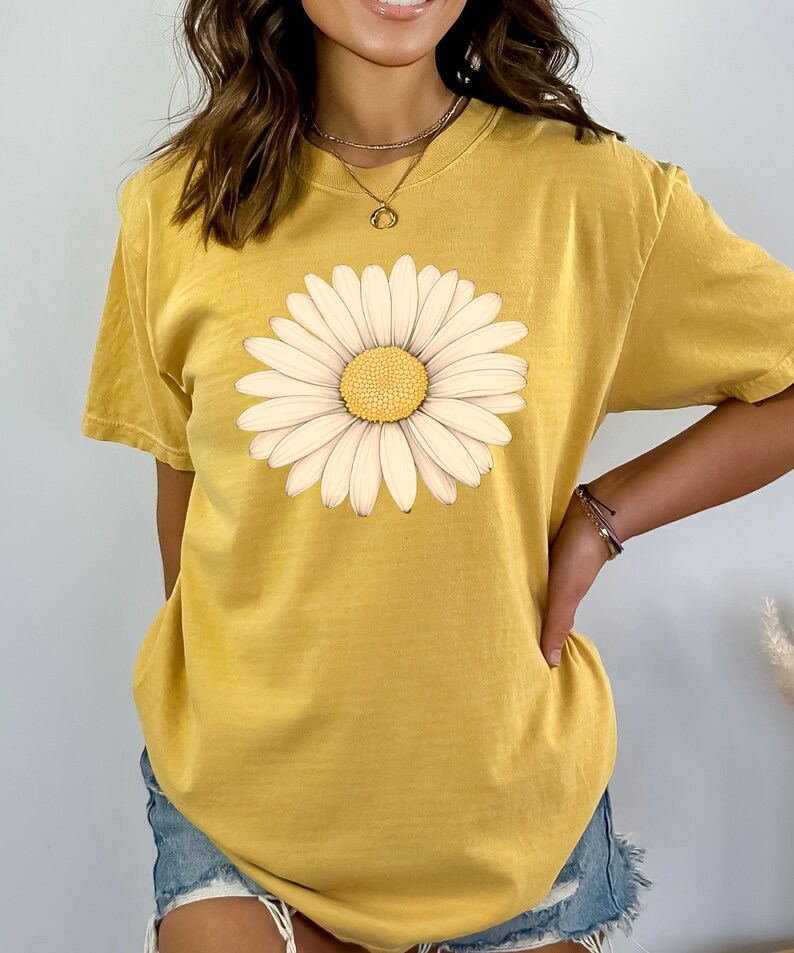Puede incluir: Una camiseta amarilla de manga corta con un estampado gr&aacute;fico de margarita blanca.