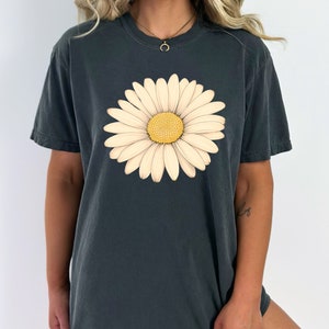 Puede incluir: Una camiseta gris oscuro con un gr&aacute;fico de margarita blanca. La margarita tiene un centro amarillo y p&eacute;talos blancos.