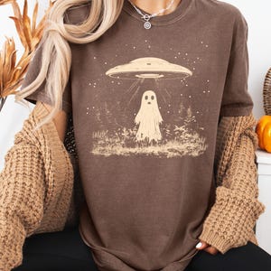 Puede incluir: Una camiseta marr&oacute;n con un gr&aacute;fico de un fantasma de pie en un bosque con un OVNI flotando por encima. El gr&aacute;fico es de estilo vintage con una gama de colores marr&oacute;n claro.