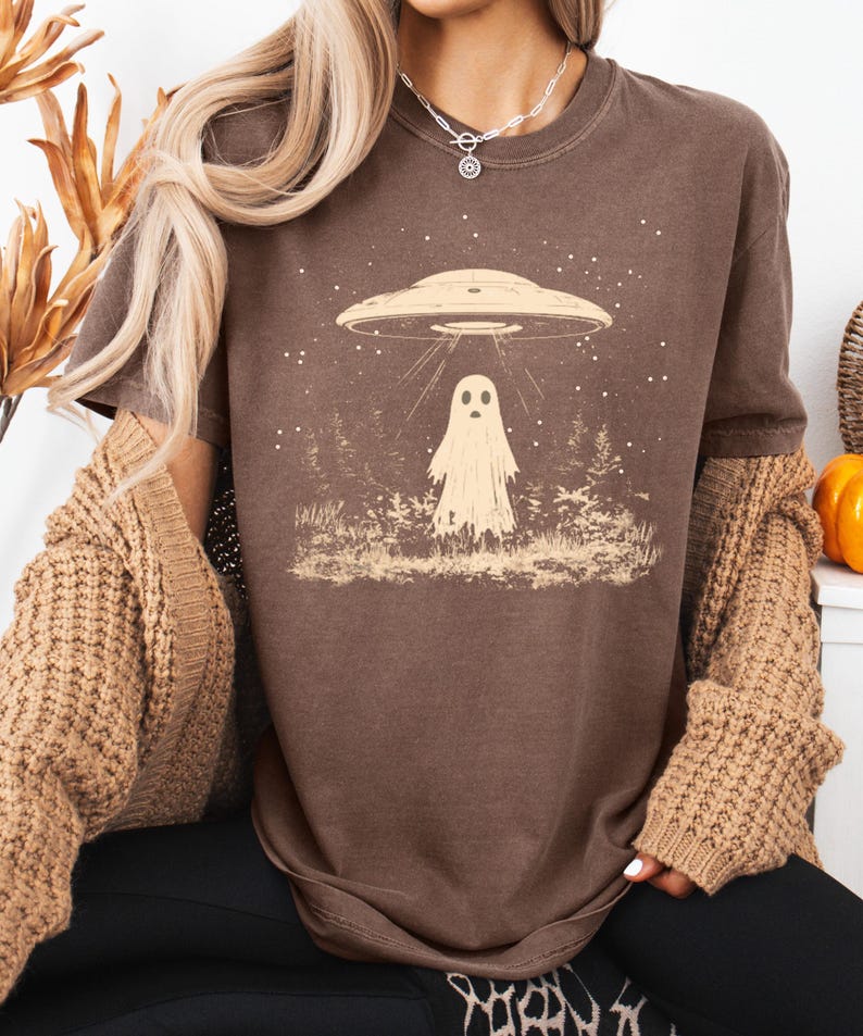 Puede incluir: Una camiseta marr&oacute;n con un gr&aacute;fico de un fantasma de pie en un bosque con un OVNI flotando por encima. El gr&aacute;fico es de estilo vintage con una gama de colores marr&oacute;n claro.