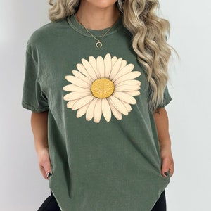 Puede incluir: Una camiseta verde con un gr&aacute;fico de margarita blanca. La margarita tiene un centro amarillo.
