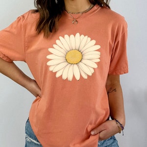 Puede incluir: Una camiseta de color coral con un gr&aacute;fico de margarita blanca. La margarita tiene un centro amarillo.