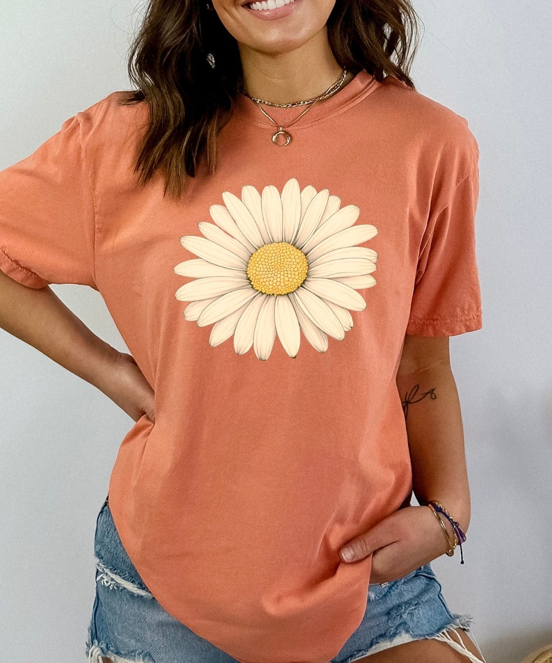 Puede incluir: Una camiseta de color coral con un gr&aacute;fico de margarita blanca. La margarita tiene un centro amarillo.