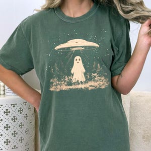 Puede incluir: Una camiseta verde con un gr&aacute;fico de un fantasma siendo abducido por un OVNI. El fantasma es blanco y el OVNI es gris. El gr&aacute;fico est&aacute; rodeado de estrellas.