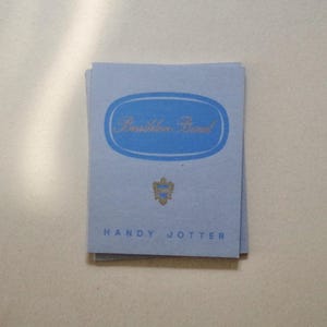 Op de afbeelding: Een stapel lichtblauwe kaarten met gouden tekst en een blauw ovaal ontwerp. De tekst "Davidson Brand" staat in het ovaal. Daaronder staat een gouden embleem en de woorden "HANDY JOTTER" staan onderaan.