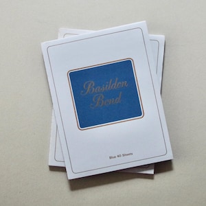 Könnte beinhalten: Ein Stapel weißer Notizblöcke mit einem blauen Quadrat und goldenem Text, der "Basildon Bond" lautet. Die Notizblöcke haben einen dünnen goldenen Rand und den Text "Blue 40 Sheets" unten.