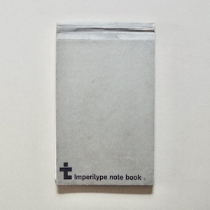 Può includere: Un piccolo taccuino rettangolare con una copertina grigio chiaro. Il taccuino ha un design semplice con la scritta "Imperitype note book" stampata in nero in basso a destra. Il taccuino è su sfondo bianco.