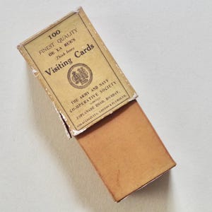 Pode incluir: Uma caixa vintage aberta de "Visiting Cards" com o texto "100 Finest Quality De La Rue's". A caixa &eacute; bege com interior castanho e texto de "The Army and Navy Co-operative Society".