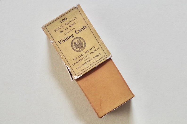 Pode incluir: Uma caixa vintage aberta de "Visiting Cards" com o texto "100 Finest Quality De La Rue's". A caixa &eacute; bege com interior castanho e texto de "The Army and Navy Co-operative Society".