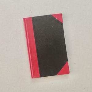 May include: A small, rectangular book with a black cover and a red spine and edges. The book's cover is divided into two sections, with the black section taking up most of the surface area. The red spine and edges provide a contrasting colour.