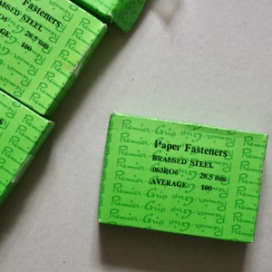 Puede incluir: Una caja verde de grapas de papel Premier Grip de acero niquelado. La caja contiene 100 grapas de 28,5 mm de longitud. La caja tiene el texto "Premier Grip Paper Fasteners, BRASSED STEEL, 063R06 28,5 mm, AVERAGE 100" impreso en ella.