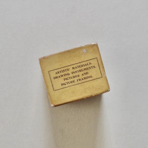 Pode incluir: Uma pequena caixa retangular vintage com o texto "ARTISTS' MATERIALS, DRAWING INSTRUMENTS, PICTURES AND PICTURE FRAMING". A caixa &eacute; de cor amarela desbotada sobre um fundo branco.