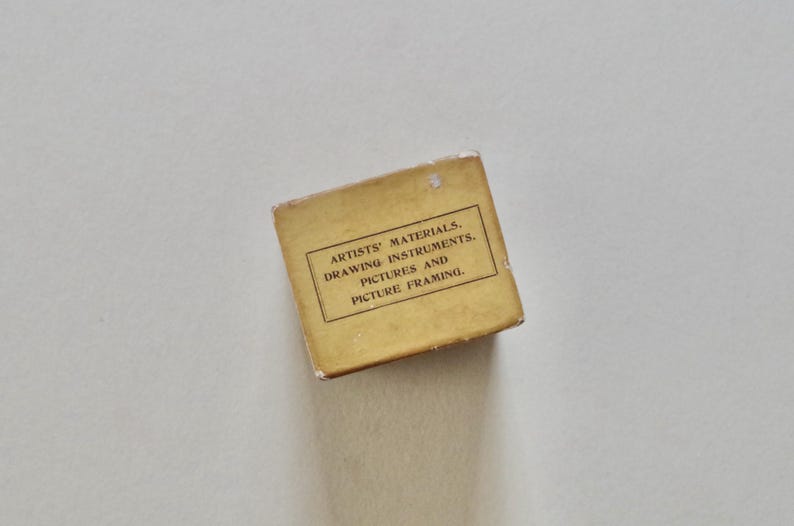 Pode incluir: Uma pequena caixa retangular vintage com o texto "ARTISTS' MATERIALS, DRAWING INSTRUMENTS, PICTURES AND PICTURE FRAMING". A caixa &eacute; de cor amarela desbotada sobre um fundo branco.